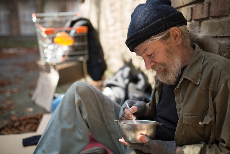 Close up view of homeless man sitting by the brick wall, eating. Homeless man with his belongings living in the street.
