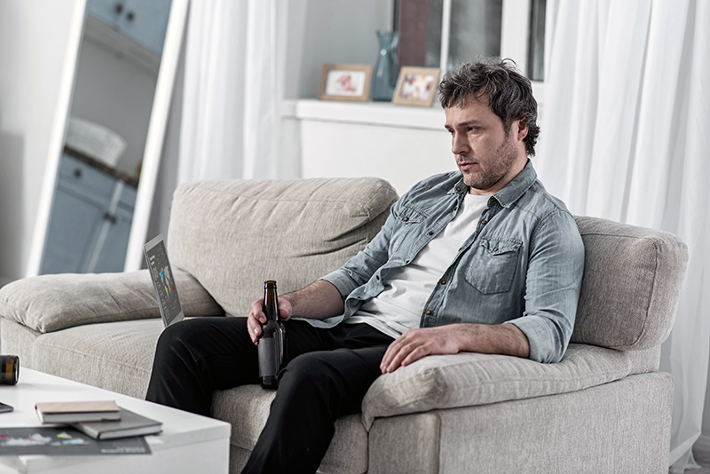 Depression. Unshaven depressed man sitting on a soft sofa with a laptop by his side and drinking alcohol after losing his job
