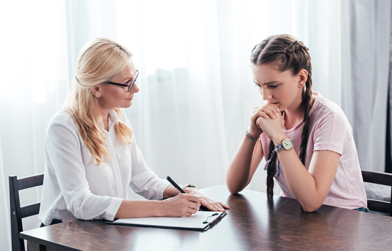stressed teenage girl sitting at table on therapy session by female counselor writing in clipboard in office